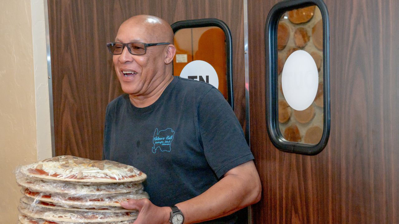 James Carson smiles as he walks into the kitchen with several pizzas at Nelsen's Hall & Bitters Club on June 15, 2026, on Washington Island, Wis. Carson works in the kitchen at the historic tavern two days per week. He bartends at the Albatross Drive-In, as well. (Heidi Hodges for Wisconsin Watch)